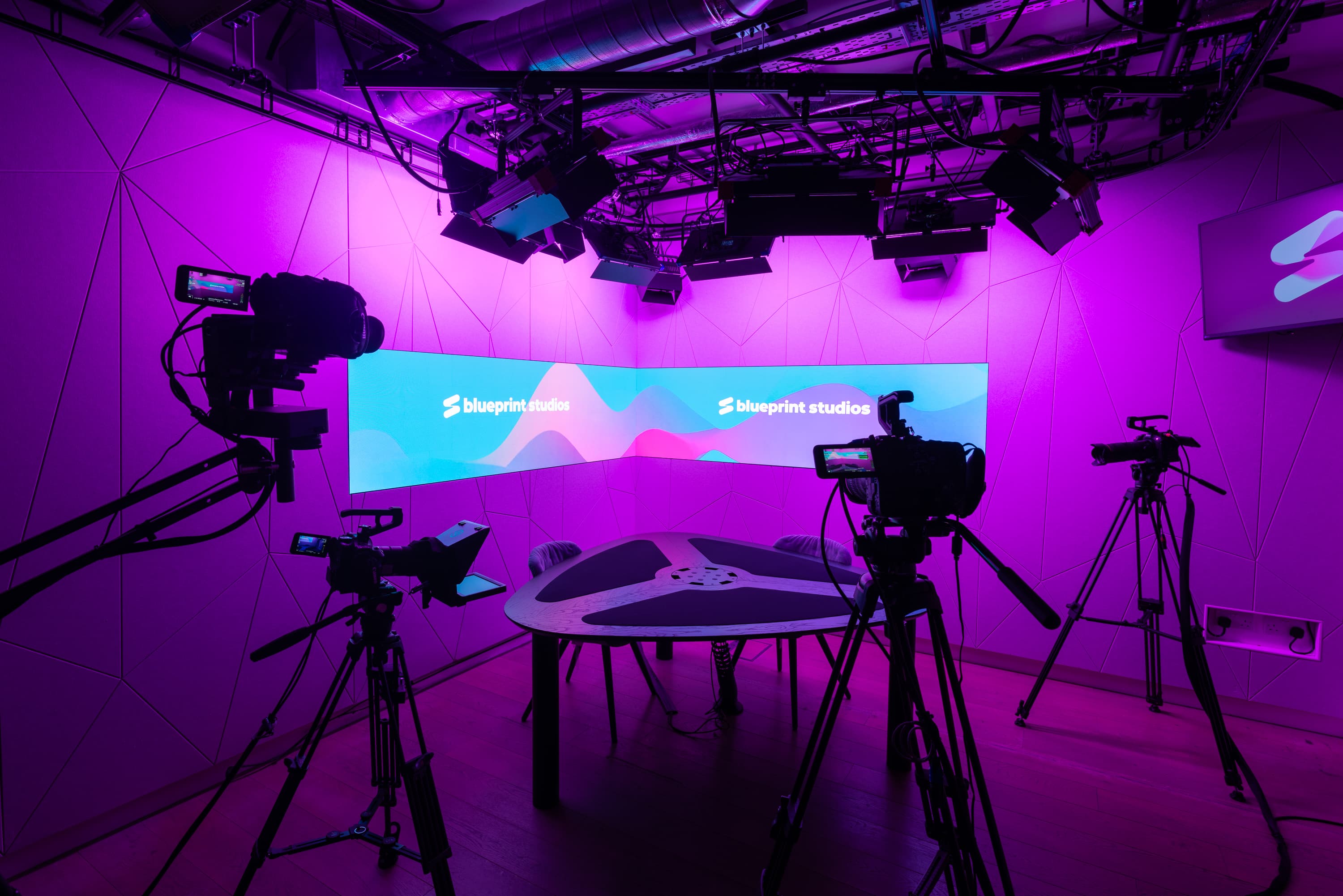 The broadcast set at Blueprint Studios London, in low purple lighting, with cameras pointing at an empty table and chairs, with an LED screen showcasing the Blueprint Studios logo