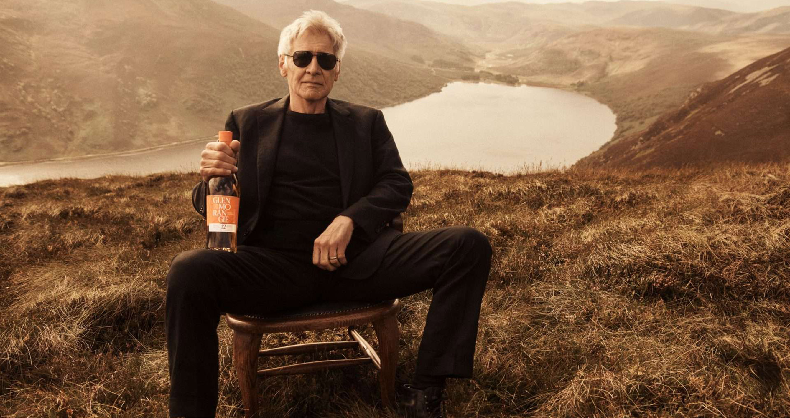 Harrison Ford, wearing black sunglasses, black blazer, black tshirt and black trousers, sitting in the mountains with a bottle of Glenmorangie