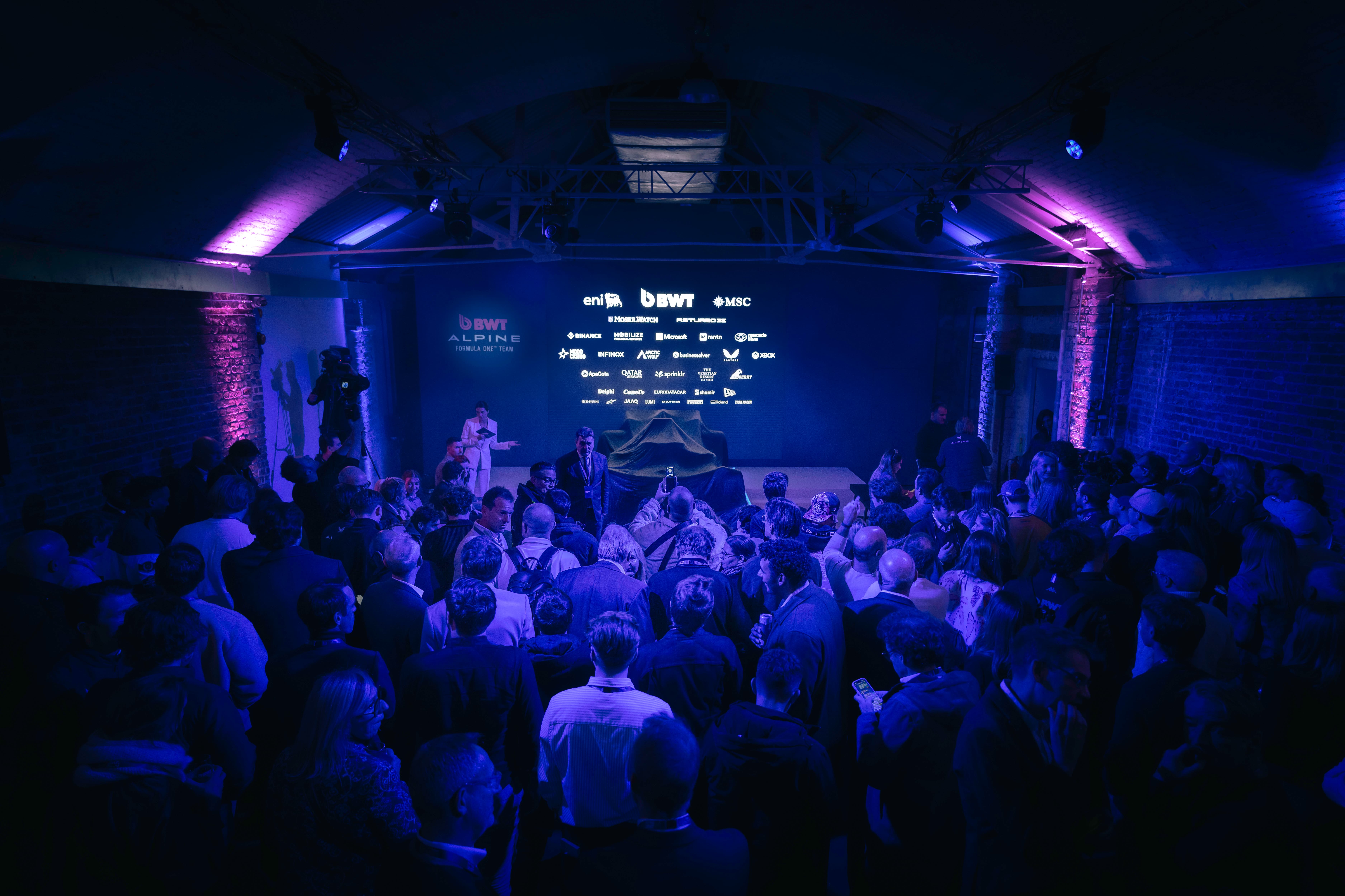 A packed room in low lighting, with people crowded in front of a stage with a Formula 1 car under cover, ready to be revealed